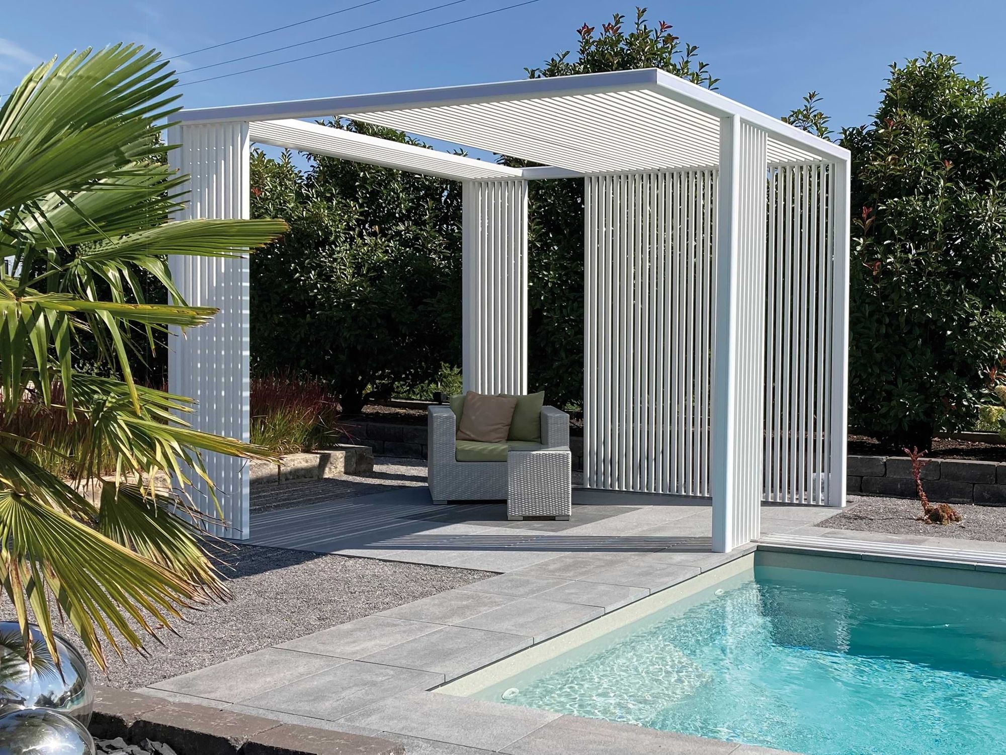 Moderne weiße Pergola mit vertikalen Lamellen, mit Blick auf einen Pool, umgeben von Grün und einer Palme, die einen ruhigen Platz im Freien bietet.