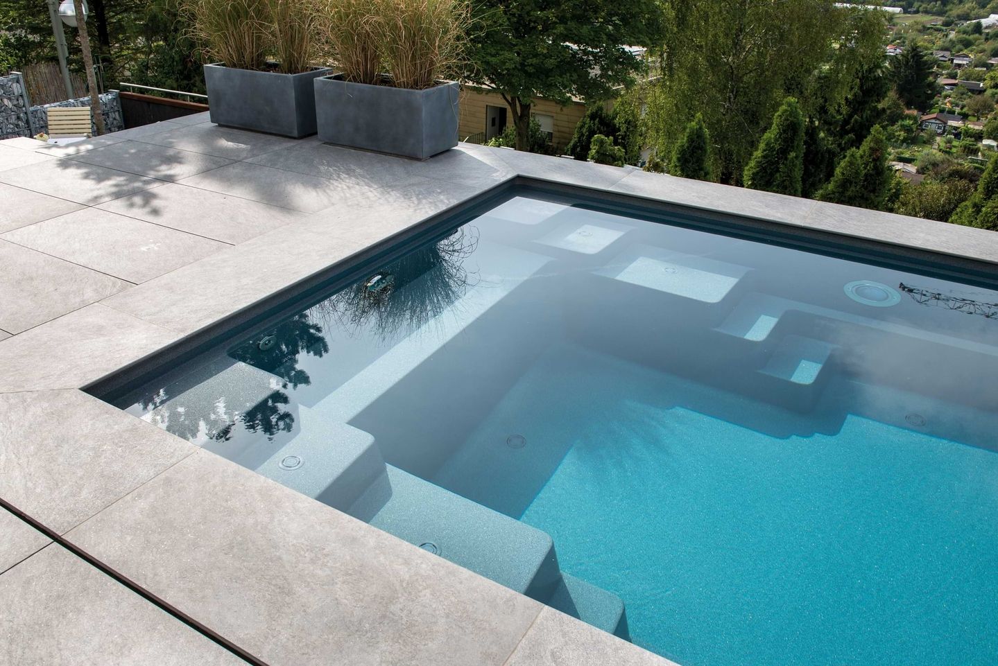 Ein ruhiger Swimmingpool mit einem glatten Steindeck, umgeben von üppigem Grün und modernen Pflanzgefäßen, in denen sich der blaue Himmel spiegelt.