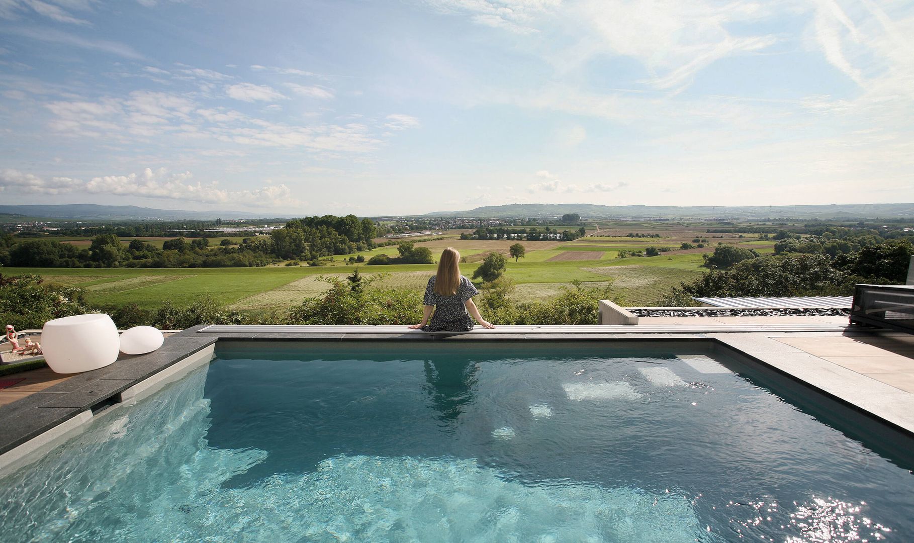 Eine Frau sitzt an einem ruhigen Pool und blickt auf saftig grüne Felder und ferne Hügel unter einem strahlend blauen Himmel.