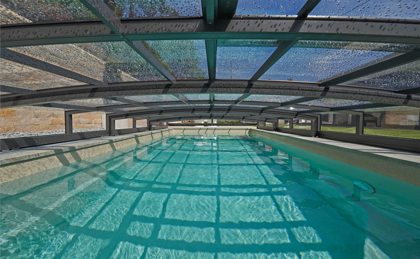 Ein klares Hallenbad unter einem gewölbten Glasdach, in dem sich das Sonnenlicht auf der Wasseroberfläche spiegelt.